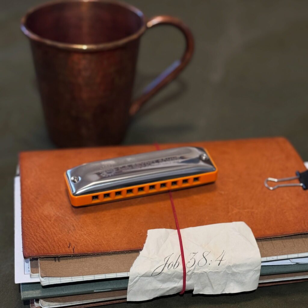 A leather-bound heirloom journal stacked with notebooks, a harmonica, and a handwritten Job 38:4 scripture note beside a rustic copper mug, styled for a timeless gifting theme.