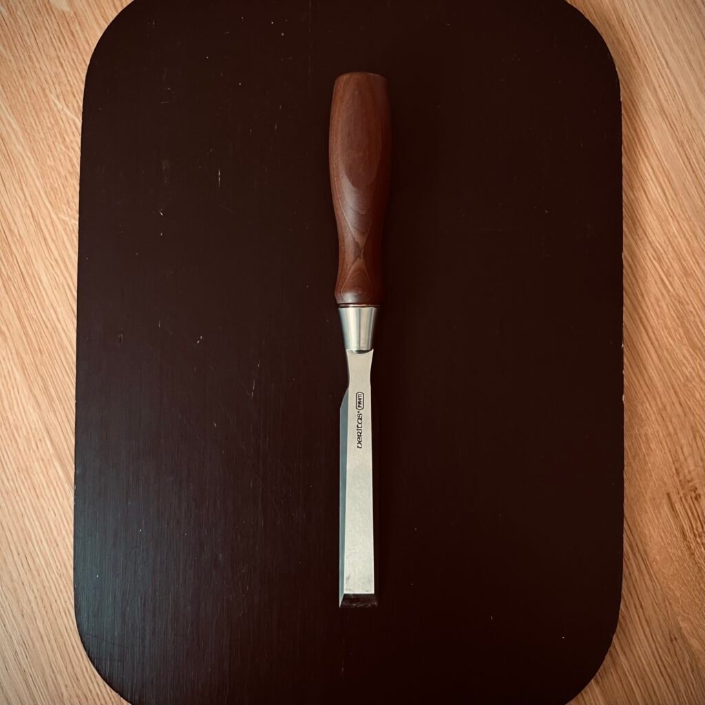 A precision woodworking chisel with a wooden handle resting on a dark wood surface, representing heirloom quality tools built for life.