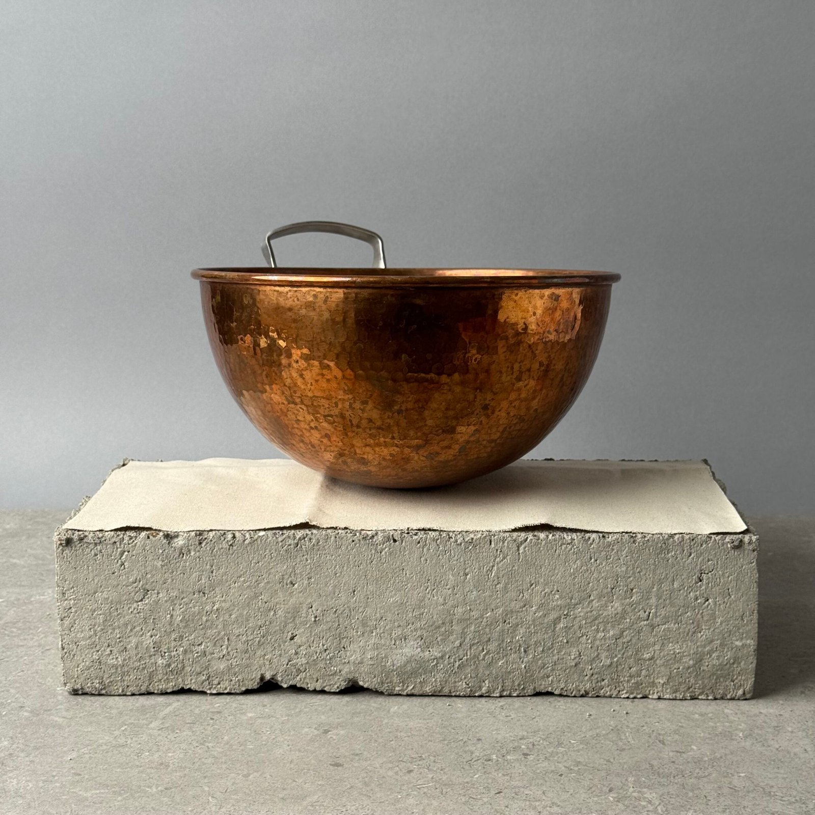 Hammered copper mixing bowl with metal handle on a concrete block against a neutral background