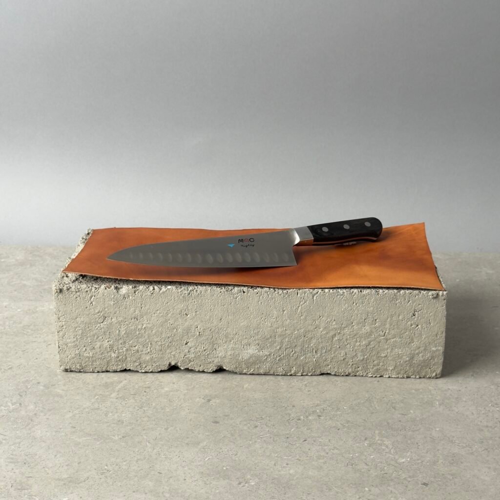 MAC MTH-80 chef knife with granton edge on a leather surface over a concrete block background